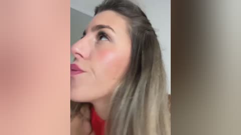 Video of a close-up of a fair-skinned woman with long, blonde hair, wearing a red top, looking upward. The background is a blurred beige wall.