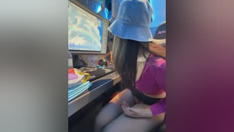 Video of a woman with long black hair wearing a blue bucket hat, sitting at a computer desk in a dimly lit room, surrounded by gaming accessories and a colorful background.