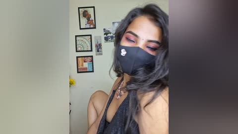 A video of a Latina woman with long black hair and dark makeup, wearing a black face mask, black tank top, and a delicate necklace, seated against a beige wall adorned with framed art and a yellow flower.