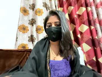 Media: Video of a South Asian woman with long black hair, wearing a black face mask, purple blouse, black cardigan, and gold necklace, seated in a room with floral-patterned curtains and brown sofa.