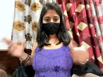 A video of a South Asian woman with medium skin tone, black hair, and a black face mask, wearing a purple lace dress, holding two glasses. Background features patterned curtains in gold, white, and maroon.