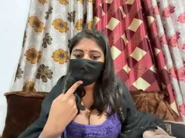 Video of a South Asian woman with medium skin tone, black hair, wearing a purple lace top, black mask, and black jacket. Background includes floral-patterned curtains and brown sofa.