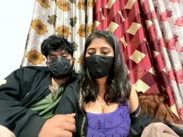 Video of a young South Asian couple in masks, wearing casual clothes, sitting on a couch. The background features floral-patterned curtains.