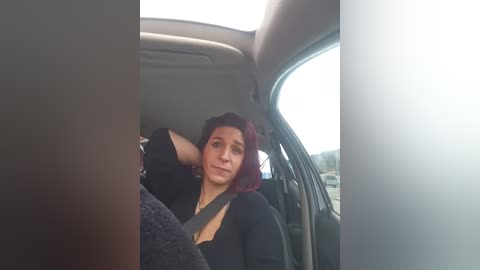 Video of a woman with shoulder-length red hair, wearing a black top, resting her head on the car seat while buckled in the driver's seat. The car interior is dimly lit with a blurry, bright background outside.