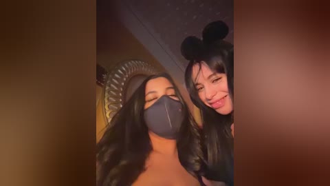 A video features two women, one with long black hair, wearing a black face mask, and the other with long black hair in a bun, smiling, in a dimly lit room.