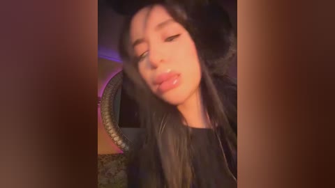 Video of an Asian woman with long black hair, wearing a black hat and jacket, leaning on a wheel, with a blurred, dimly lit background.