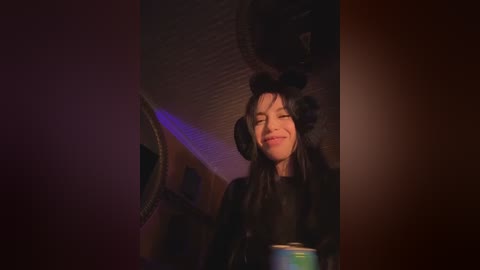 A video of a smiling woman with long dark hair, wearing black furry cat ears and a black coat, in a dimly lit room with purple lighting and a ceiling fan.