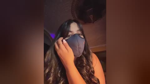 Video of a woman with long, wavy dark hair, wearing a blue mask and black top, adjusting the mask, in a dimly lit room with a ceiling fan.