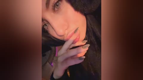 Video of a close-up of a woman's face with fair skin, blue eyes, and black hair. She has her hand to her mouth, revealing long, metallic silver nails. The background is dark and blurred, focusing attention on her expression.