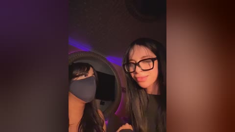 Video of a young Asian woman with long black hair, wearing black-framed glasses, a dark mask, and a black top, standing in front of a mirror reflecting her back. The background is dimly lit with purple and blue LED lights.