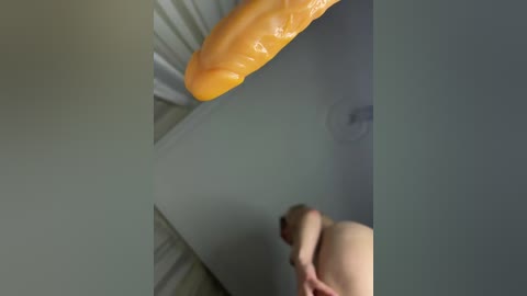 Video of a nude person with light skin, sitting on a bed with a large, realistic yellow dildo, and a blurred ceiling fan in the background.