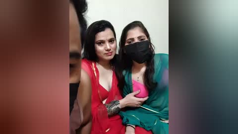 Video of two South Asian women with dark hair and medium skin tones, wearing red and green sarees, black masks, and silver jewelry, standing indoors.