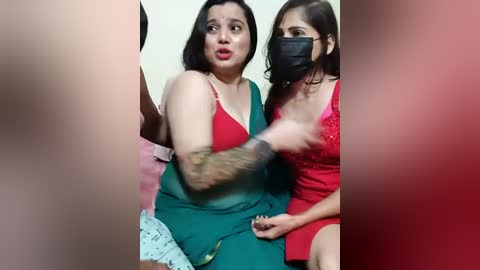 Video of a woman with long black hair, red lipstick, and a black face mask, wearing a green dress, holding a tattooed arm.