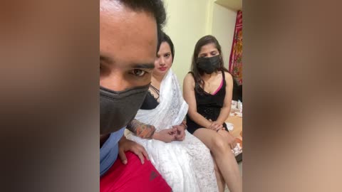 A video shows three South Asian women, two masked, sitting on a bed, one with visible tattoos. Background includes a colorful wall hanging and a wooden table.