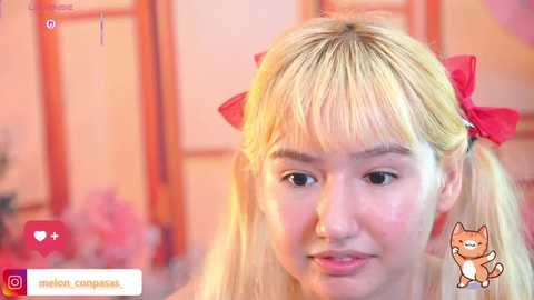 Video of an Asian woman with straight, blonde hair styled in pigtails with red bows, wearing makeup, and smiling softly. Background features blurred, warm-toned interior.