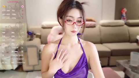Video of an Asian woman with fair skin, glasses, and purple halter top, standing in a modern living room with beige couches and decor.