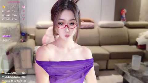 Video of an East Asian woman with fair skin, wearing red-framed glasses and a purple off-shoulder top, standing in a modern living room with beige furniture.