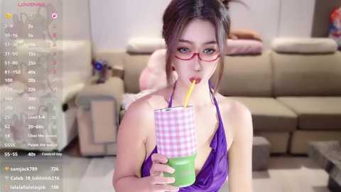 Video of an East Asian woman with light skin, brown hair, and red-framed glasses, wearing a purple halter top, drinking from a pink-and-green straw. She is in a modern living room with beige furniture.
