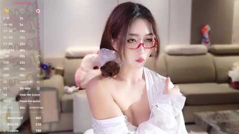 Media: Video of an East Asian woman with long dark hair, red glasses, and white lingerie, sitting on a beige couch in a modern living room.