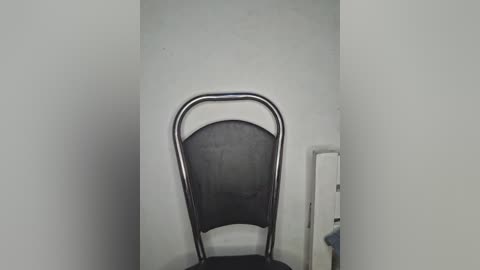 Video of a narrow, empty, white-walled corridor with a single black mesh-backed chair with metal arms against the right wall. The lighting is dim, creating a somber, minimalist atmosphere.
