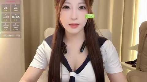 Video of an Asian woman with long brown hair, wearing a white sailor-style school uniform with a deep V-neck, sitting indoors with beige curtains in the background.