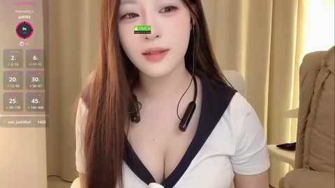 A video of a young Asian woman with long, straight brown hair, wearing a white sailor-style top with a deep V-neck, black shoulder straps, and headphones. She has fair skin and a slight smile. The background features beige curtains and a digital clock.