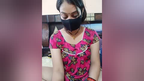 Video of a woman with medium skin tone, wearing a pink floral blouse, black face mask, and black gloves, sitting indoors with a wooden headboard and bed behind her.