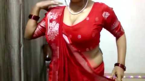 Video of a woman in a red saree with floral embroidery, wearing a pearl necklace and matching bracelets, standing indoors.