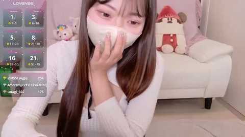 A video of a young Asian woman with long, straight, dark hair, wearing a white knitted sweater, covering her mouth with a white bandage. The background features a plush bed with stuffed toys and a pink wall.