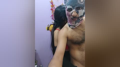Media: Video of a woman with long black hair in a red bra, embracing a shirtless man wearing a colorful, face-covering mask with a mustache. Background shows a lavender wall and festive decorations.