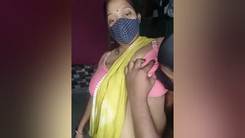 Video of a woman in a yellow saree with a blue polka-dot mask, lifting her pink bra, revealing her breasts, in a dimly-lit room with a checkered wall and a pink wall in the background.