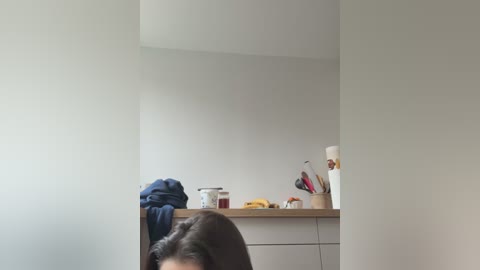 Video of a minimalist kitchen with a wooden countertop holding a white mug, utensils, and a bowl of fruit. A person with dark hair partially visible, wearing a blue shirt, leans forward.