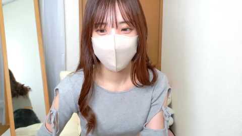Video of an Asian woman with long brown hair, wearing a light grey off-shoulder top and a white face mask, standing indoors near a wooden door and mirror.