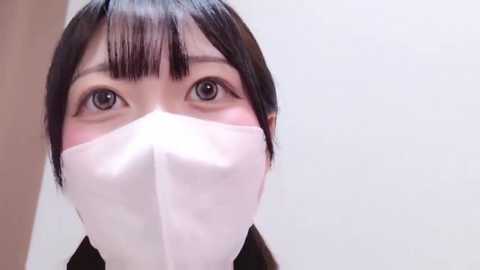 Media: Video of a woman with East Asian features, wearing a white surgical mask, black hair with blunt bangs, and light skin, standing against a plain white background.