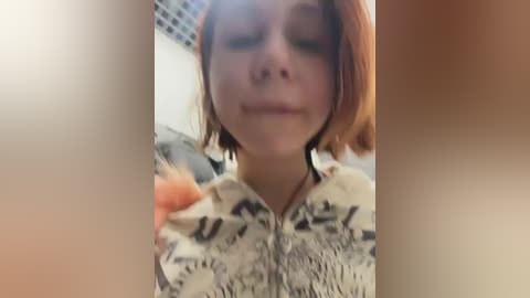 Video of a young person with reddish hair, wearing a patterned shirt, looking down with a slight smile. Background shows a blurred, indoor setting with a grid pattern.