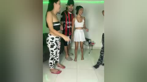 Video of three people in a modern, brightly lit room with white tiled floor and green LED strip lighting. Two women in patterned leggings and tank tops, and one man in black and red soccer jersey and shorts.