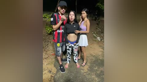 Video of three people standing outdoors: a man in a black shirt and red soccer jersey, a woman in a black top and patterned leggings, and another woman in a purple top and white skirt. Background includes dirt path and greenery.