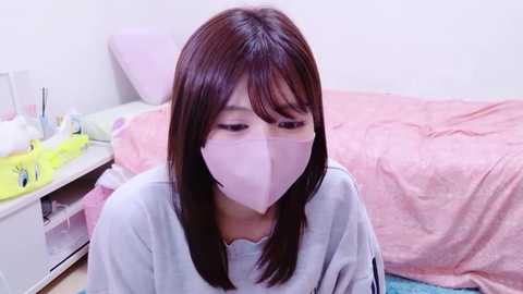 Video of a young woman with shoulder-length brown hair, wearing a light gray sweatshirt, light pink surgical mask, and a pink blanket in a pastel-colored bedroom with a bed and nightstand.