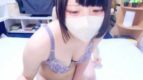Video of an Asian woman with a light skin tone, wearing a lavender lace bra and a white mask, kneeling on a white surface, with a cluttered room in the background.