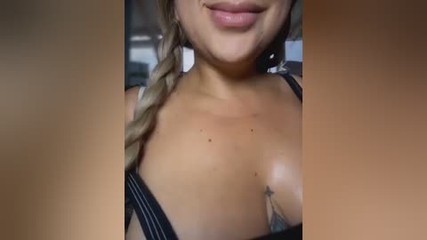 Video of a smiling woman with light skin, braided blonde hair, wearing a black lace bra, showcasing her medium-sized breasts. Background is blurred, indoor setting with neutral tones.