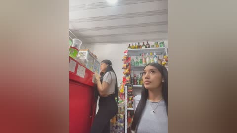 Video of a convenience store with a young woman in a gray shirt and black pants examining items in a red fridge, while another woman in a white shirt and black pants looks on.