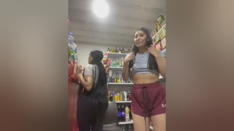 Video of two young women in a convenience store: one in a black outfit, the other in a grey crop top and maroon shorts. Shelves filled with beverages and snacks in the background.