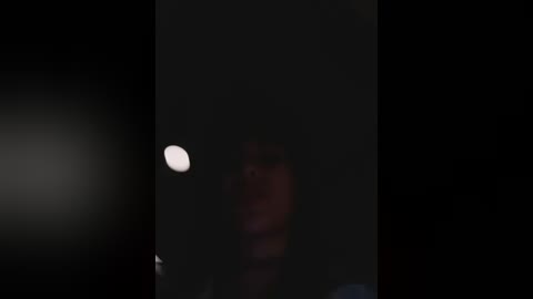 A dimly lit video captures a woman with a blurred face, possibly Asian, wearing a dark top and visible hair, standing against a dark, shadowy background. A faint light source, possibly a moon, is visible in the upper left corner.