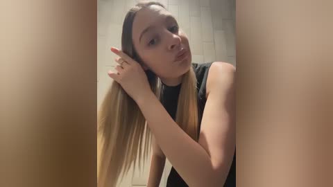 Media: Video of a young woman with long, straight blonde hair, light skin, and a slender physique, wearing a black top, looking pensively into the camera while in a bathroom with white tiled walls.