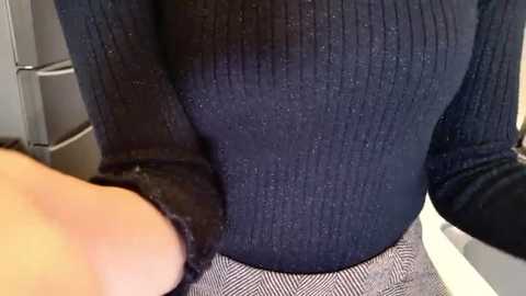 Video of a person's hand grasping a dark blue, ribbed-knit sweater. The sweater is snug-fitting, highlighting a curvaceous figure. Background features a blurred, modern kitchen with stainless steel appliances and a white countertop.