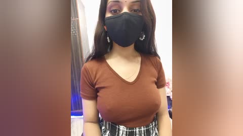 Video of a young woman with medium skin tone, wearing a black face mask, a brown V-neck t-shirt, and a plaid skirt, in a blurred indoor setting.