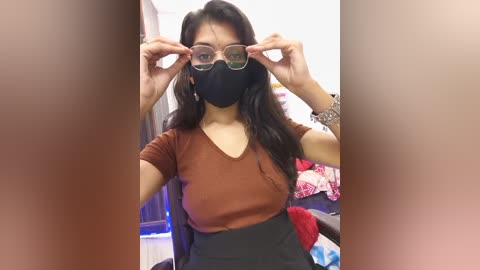 Video of a South Asian woman with long black hair, wearing a black mask, clear glasses, a rust-colored top, and a black skirt, adjusting her glasses in a cluttered room with red and white patterned clothing.