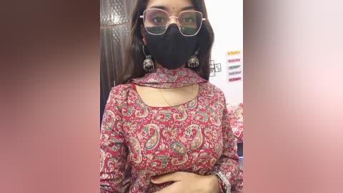 Video of a South Asian woman with long black hair, wearing a red paisley-patterned top, black face mask, and clear glasses, standing indoors.