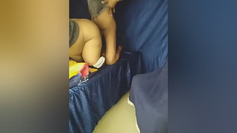 Video of a nude woman with medium brown skin, lying face down on a blue bedspread, wearing only a black thong, with her buttocks prominently visible.