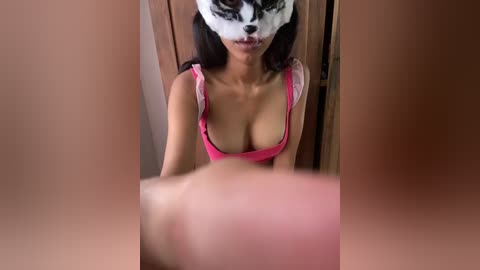 Video of a woman with medium brown skin and long black hair, wearing a pink bra, with a raccoon mask covering her face.